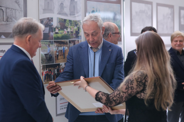 Dwóch mężczyzn i kobieta w galerii sztuki. Mężczyzna w garniturze wręcza ramę z obrazem.