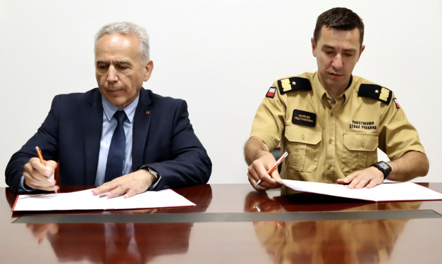 Dwóch mężczyzn przy stole podpisuje dokumenty. Jeden w garniturze, drugi w mundurze Państwowej Straży Pożarnej.