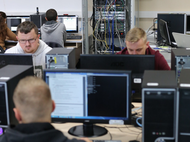 Ludzie pracują przy komputerach w sali lekcyjnej. Kluczowe obiekty: monitory, komputery, szafa serwerowa.