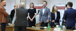 Grupa osób stoi przy drewnianym stole, na którym znajdują się kubki, termosy i butelka wody.