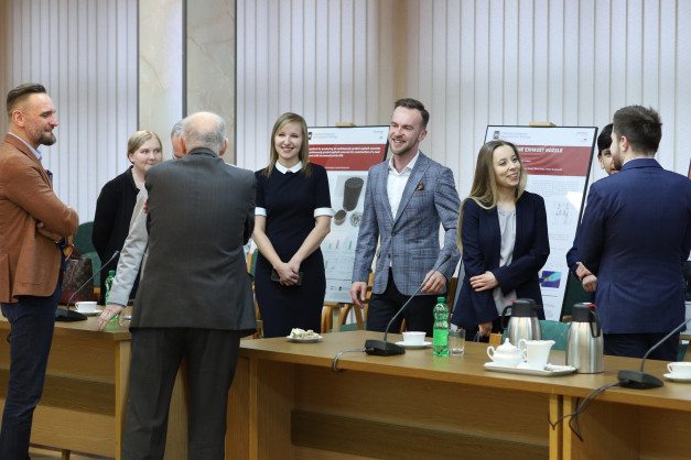 Grupa osób stoi przy drewnianym stole, na którym znajdują się kubki, termosy i butelka wody.