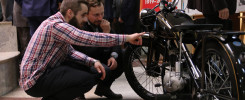 Dwóch mężczyzn ogląda zabytkowy motocykl. W tle widoczny baner z datą 1919-2019.