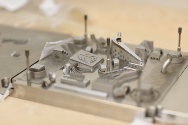 Metalowe wydruki 3D, w tym elementy zębate i kształty geometryczne, na metalowej płycie.