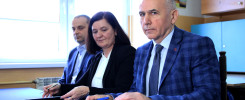 Trzy osoby siedzą przy stole, jedna z nich podpisuje dokument. W tle widać okno i tablicę z kompasem.