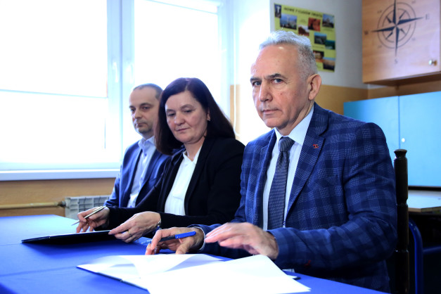 Trzy osoby siedzą przy stole, jedna z nich podpisuje dokument. W tle widać okno i tablicę z kompasem.