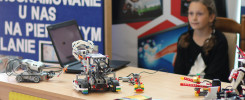 Robotyka i programowanie na pierwszym planie. Na stole roboty z klocków, laptop, książki. Dziewczynka w tle.
