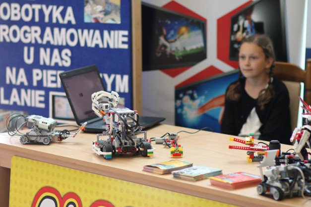Robotyka i programowanie na pierwszym planie. Na stole roboty z klocków, laptop, książki. Dziewczynka w tle.