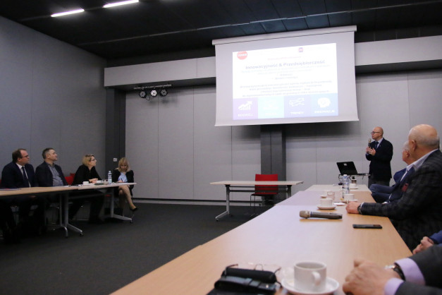 Konferencja z prezentacją na ekranie. Uczestnicy siedzą przy stołach, jeden mężczyzna przemawia.