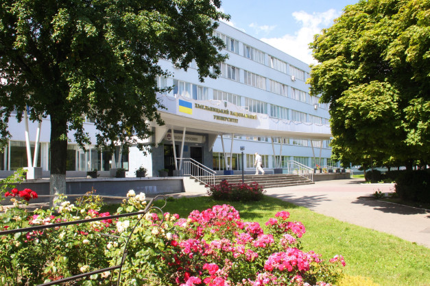 Budynek uniwersytetu z flagą Ukrainy. Kluczowe obiekty: budynek, drzewa, kwiaty, schody, osoba. Kontekst: teren uczelni.