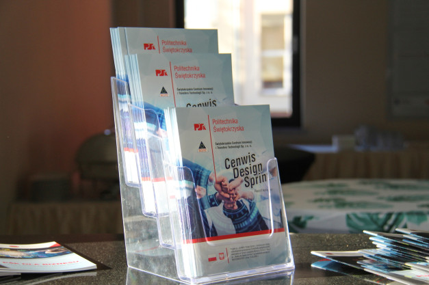 Ulotki Politechniki Świętokrzyskiej w przezroczystym stojaku. Widoczne logo uczelni i tytuł "Cenwis Design Sprint".
