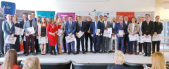Grupa osób w strojach formalnych stoi na scenie, trzymając certyfikaty. W tle widoczne są banery.