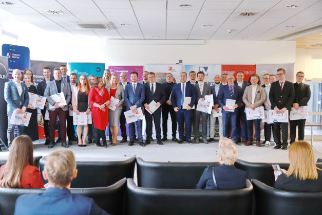 Grupa osób w strojach formalnych stoi na scenie, trzymając certyfikaty. W tle widoczne są banery.