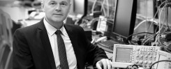 Mężczyzna w garniturze siedzi przy biurku z aparaturą elektroniczną. W tle monitory i sprzęt laboratoryjny.