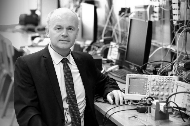 Mężczyzna w garniturze siedzi przy biurku z aparaturą elektroniczną. W tle monitory i sprzęt laboratoryjny.