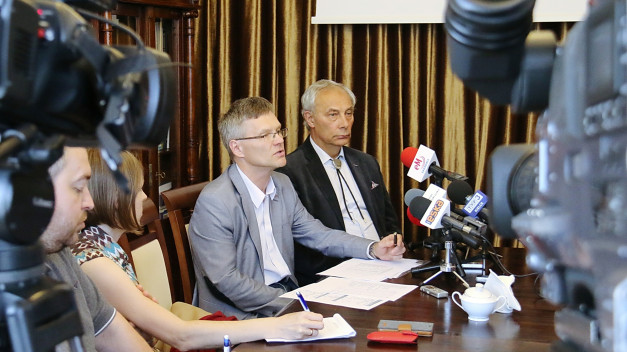 Konferencja prasowa z udziałem mężczyzn przy stole, otoczona przez kamery i mikrofony.