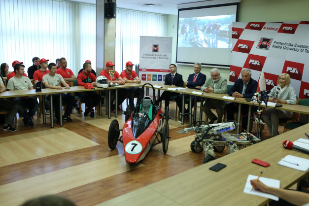 Grupa osób siedzi przy stole, przed nimi czerwony bolid wyścigowy i robot. Kontekst: wydarzenie Politechniki Świętokrzyskiej.