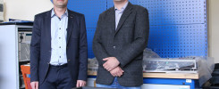Dwóch mężczyzn w garniturach stoi przed niebieską tablicą perforowaną. W tle widać sprzęt elektroniczny i głośnik.