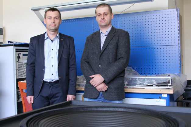 Dwóch mężczyzn w garniturach stoi przed niebieską tablicą perforowaną. W tle widać sprzęt elektroniczny i głośnik.