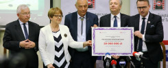 Cztery osoby stoją za zielonym stołem, trzymając certyfikat. Kluczowe obiekty: certyfikat, mikrofony, ludzie.