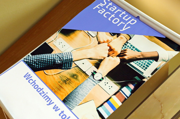 Ręce zaciśnięte w pięści na tle laptopa i listew zasilających. Napisy "Startup Factory" i "Wchodzimy w to".