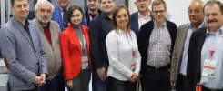 Grupa osób w strojach biznesowych pozuje do zdjęcia w pomieszczeniu z wyposażeniem technicznym.