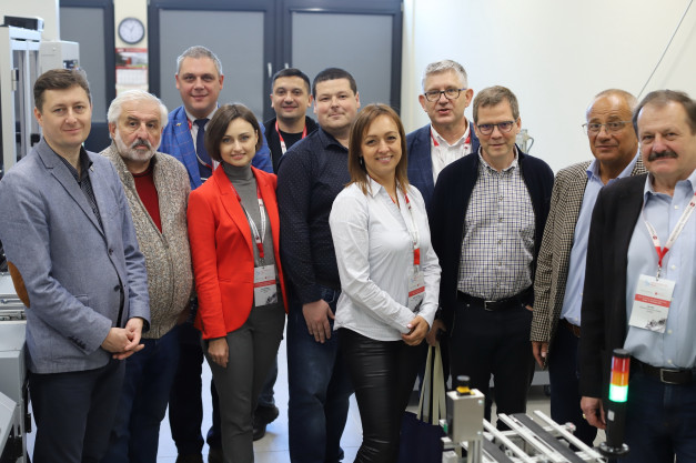 Grupa osób w strojach biznesowych pozuje do zdjęcia w pomieszczeniu z wyposażeniem technicznym.