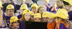Dzieci w żółtych czapkach patrzą na laptopa. Kluczowe obiekty: dzieci, laptop, czapki. Kontekst: wydarzenie edukacyjne.