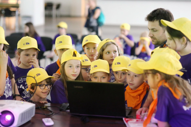 Dzieci w żółtych czapkach patrzą na laptopa. Kluczowe obiekty: dzieci, laptop, czapki. Kontekst: wydarzenie edukacyjne.