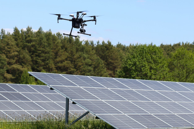 Dron lata nad panele słoneczne. Tło stanowi las i błękitne niebo.