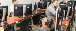 Ludzie siedzą przy komputerach w sali lekcyjnej. Kluczowe obiekty: monitory Dell, laptopy, krzesła, biurka.
