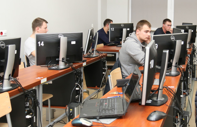 Ludzie siedzą przy komputerach w sali lekcyjnej. Kluczowe obiekty: monitory Dell, laptopy, krzesła, biurka.