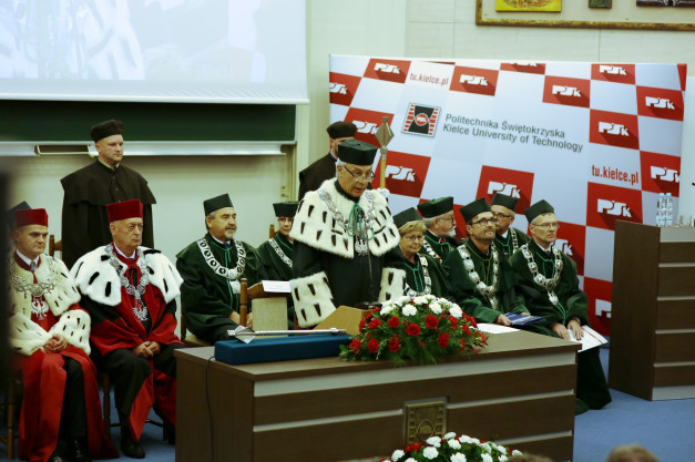 Uroczystość akademicka z udziałem osób w togach i łańcuchach. Kluczowe obiekty: togi, łańcuchy, podium, kwiaty.