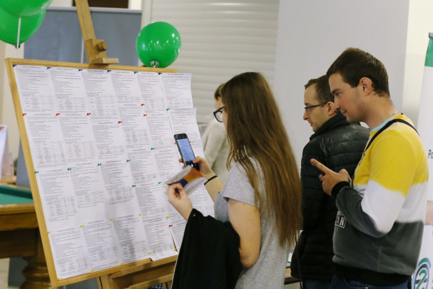 Osoby przy tablicy z ogłoszeniami, jedna z nich robi zdjęcie telefonem. Kontekst: targi pracy lub wydarzenie informacyjne.