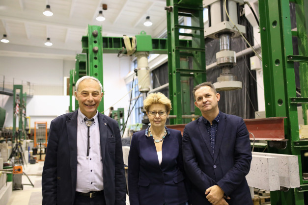 Trzy osoby stoją przed maszynami w laboratorium. Widać betonowe elementy i zielone konstrukcje.