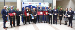 Grupa osób w garniturach i sukienkach odbiera nagrody. Kluczowe obiekty: nagrody, róże, baner.