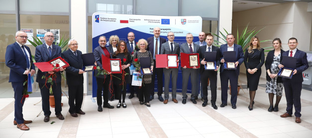 Grupa osób w garniturach i sukienkach odbiera nagrody. Kluczowe obiekty: nagrody, róże, baner.