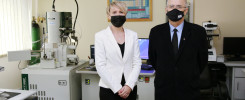 Dwie osoby w maseczkach stoją w laboratorium obok mikroskopu elektronowego. W tle widać tablicę z układem okresowym.