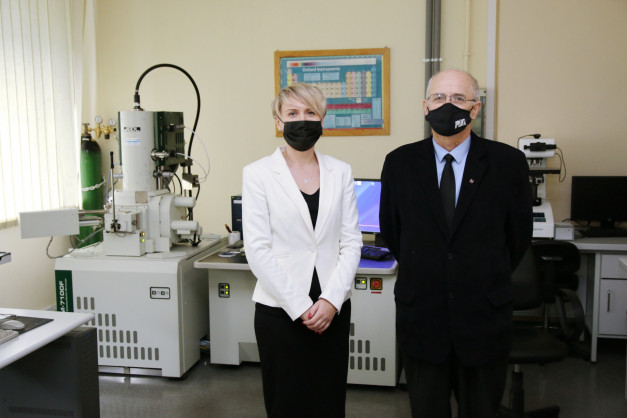 Dwie osoby w maseczkach stoją w laboratorium obok mikroskopu elektronowego. W tle widać tablicę z układem okresowym.
