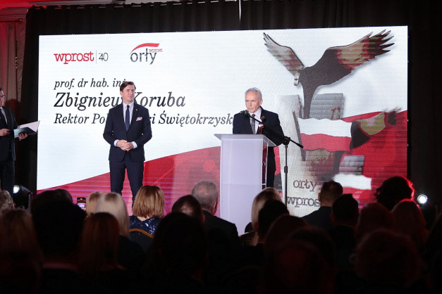 Dwóch mężczyzn stoi na scenie przed ekranem z logo "wprost" i orłem. Jeden z nich przemawia przy mównicy.