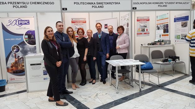 Grupa osób stoi przed stoiskami wystawowymi. W tle widoczne są banery i plakaty.