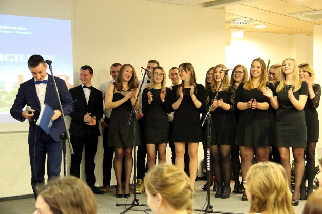 Grupa młodych ludzi w strojach galowych stoi na scenie z mikrofonami. W tle ekran projekcyjny.