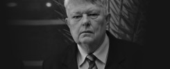 Czarno-białe zdjęcie portretowe starszego mężczyzny w garniturze i krawacie. Tło stanowi ciemna ściana z rośliną.