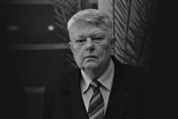 Czarno-białe zdjęcie portretowe starszego mężczyzny w garniturze i krawacie. Tło stanowi ciemna ściana z rośliną.