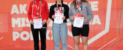 Trzy kobiety na podium z dyplomami i medalami. Kluczowe obiekty: kobiety, dyplomy, medale, podium.