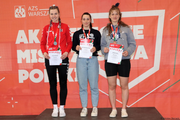Trzy kobiety na podium z dyplomami i medalami. Kluczowe obiekty: kobiety, dyplomy, medale, podium.