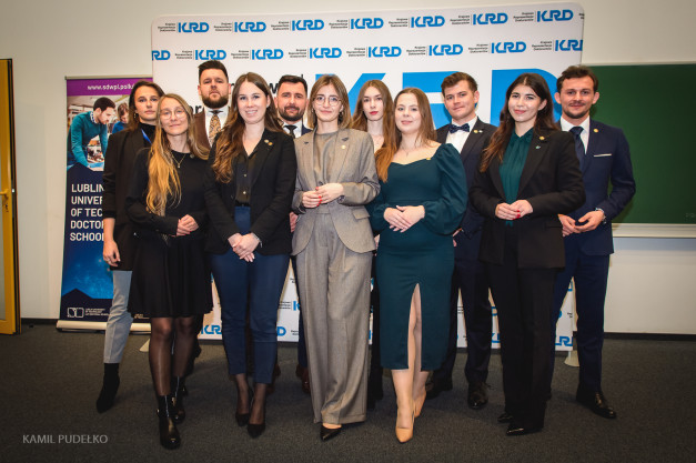 Grupa młodych ludzi pozuje do zdjęcia przed banerem KRD. Wszyscy ubrani są formalnie.