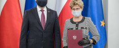 Mężczyzna i kobieta w maseczkach stoją przed flagami Polski i UE. Kobieta trzyma dokument z godłem Polski.