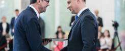 Dwóch mężczyzn w garniturach ściska sobie dłonie. Jeden z nich wręcza drugiemu dokument. W tle widoczni są ludzie.
