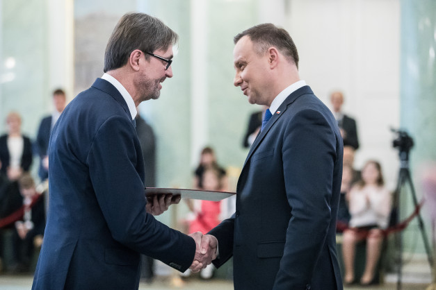Dwóch mężczyzn w garniturach ściska sobie dłonie. Jeden z nich wręcza drugiemu dokument. W tle widoczni są ludzie.