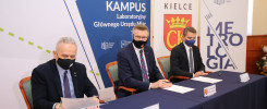 Trzech mężczyzn w garniturach i maskach siedzi przy stole. Na stole dokumenty i długopisy. Tło to banery z logo instytucji.
